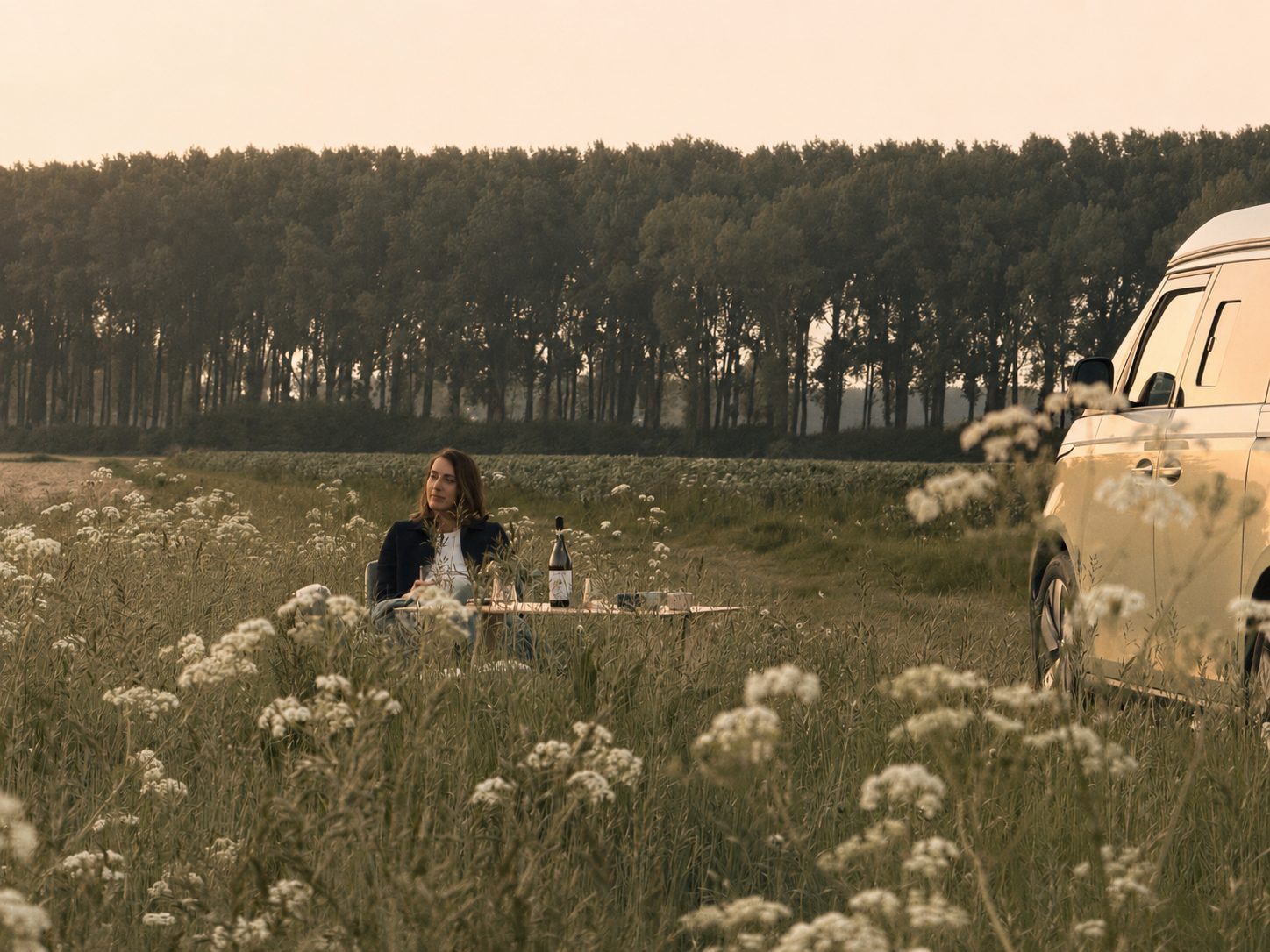 A woman at a table beside the van in a flowering meadow at golden hour